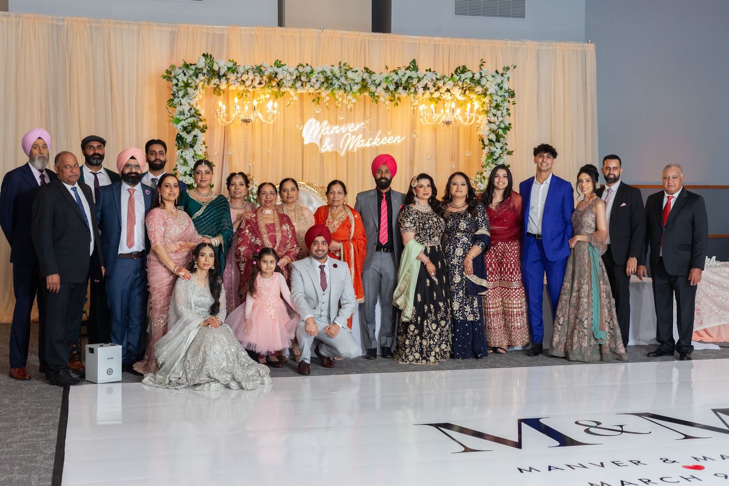 Large wedding party in colorful traditional and formal attire poses on a dance floor in front of a floral backdrop at an indoor reception.
