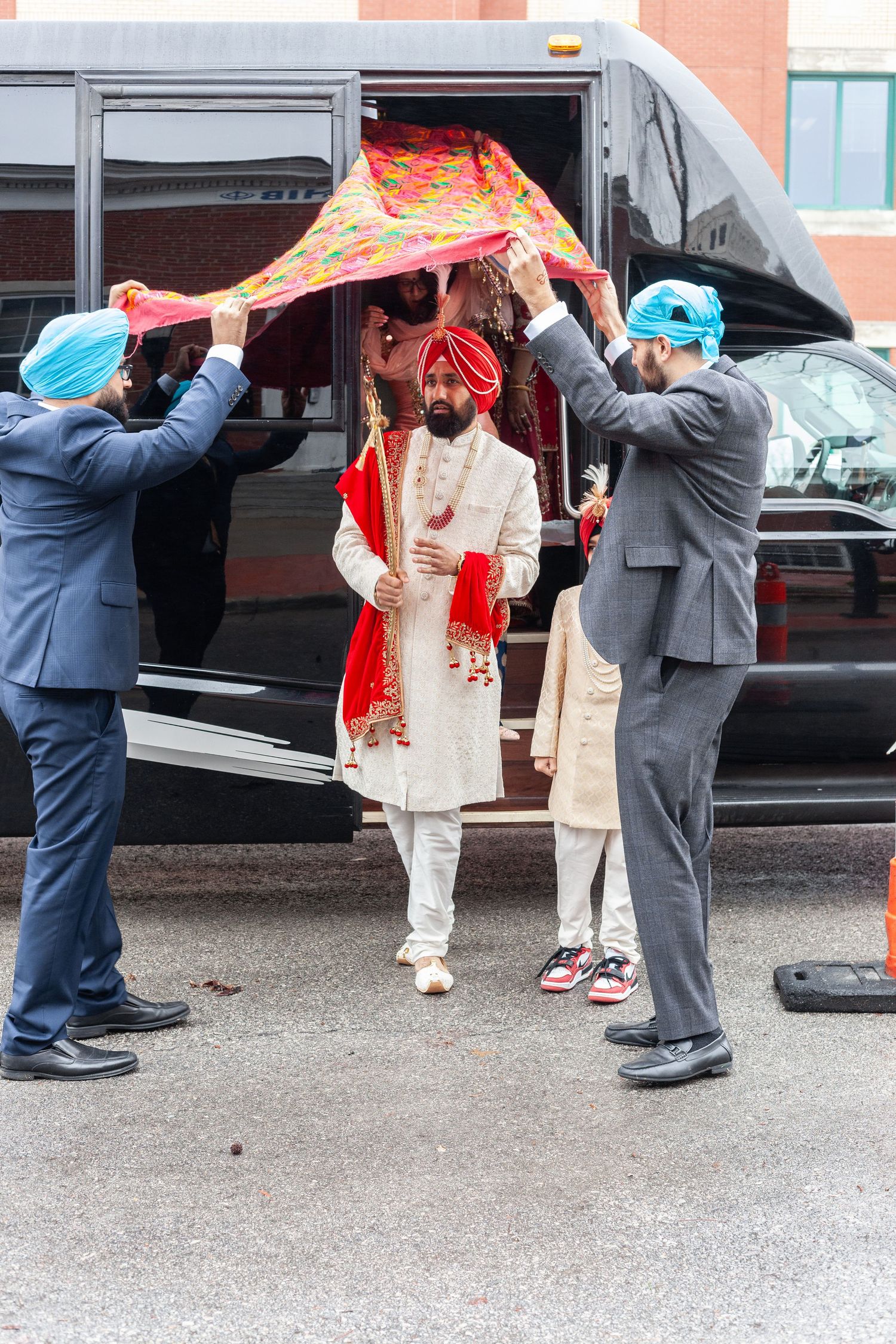 Groom in a white sherwani and red turban walks under a colorful phulkari canopy held by men in blue turbans at a Sikh wedding.