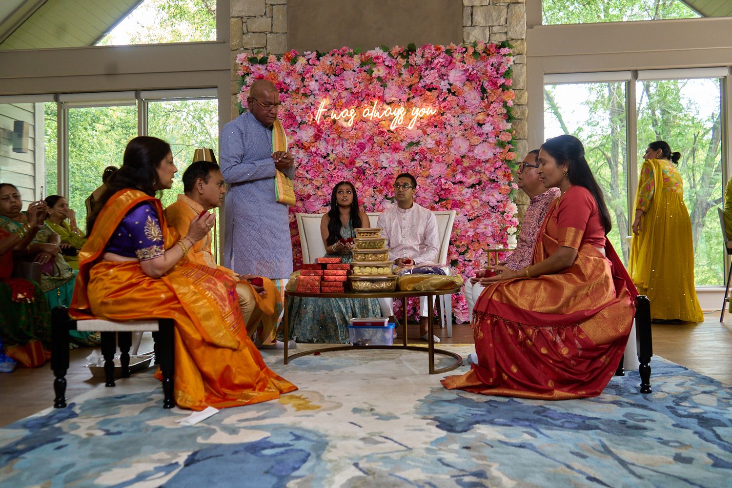 Women in colorful traditional Indian saris gather around a ceremonial table in front of a pink floral wall at an Indian wedding ceremony.