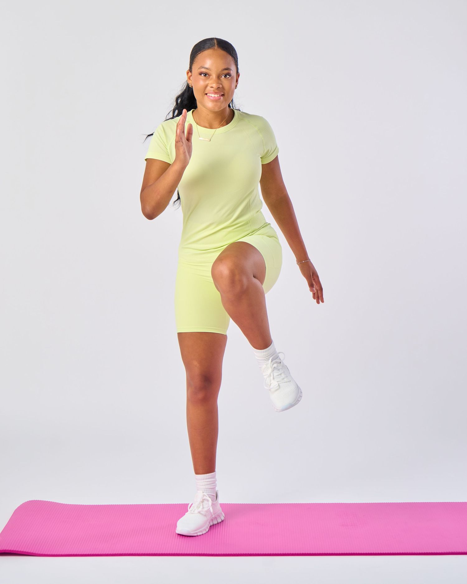 Woman in lime green athletic t-shirt and bike shorts poses on pink yoga mat wearing white sneakers.