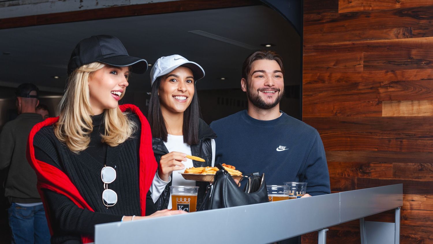 Three friends smiling and enjoying drinks and snacks at an outdoor bar or restaurant venue.