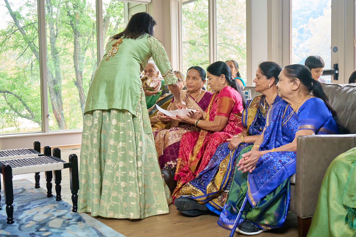 Women in colorful traditional Indian saris and a green outfit gather in a bright sunlit living room during a cultural ceremony.