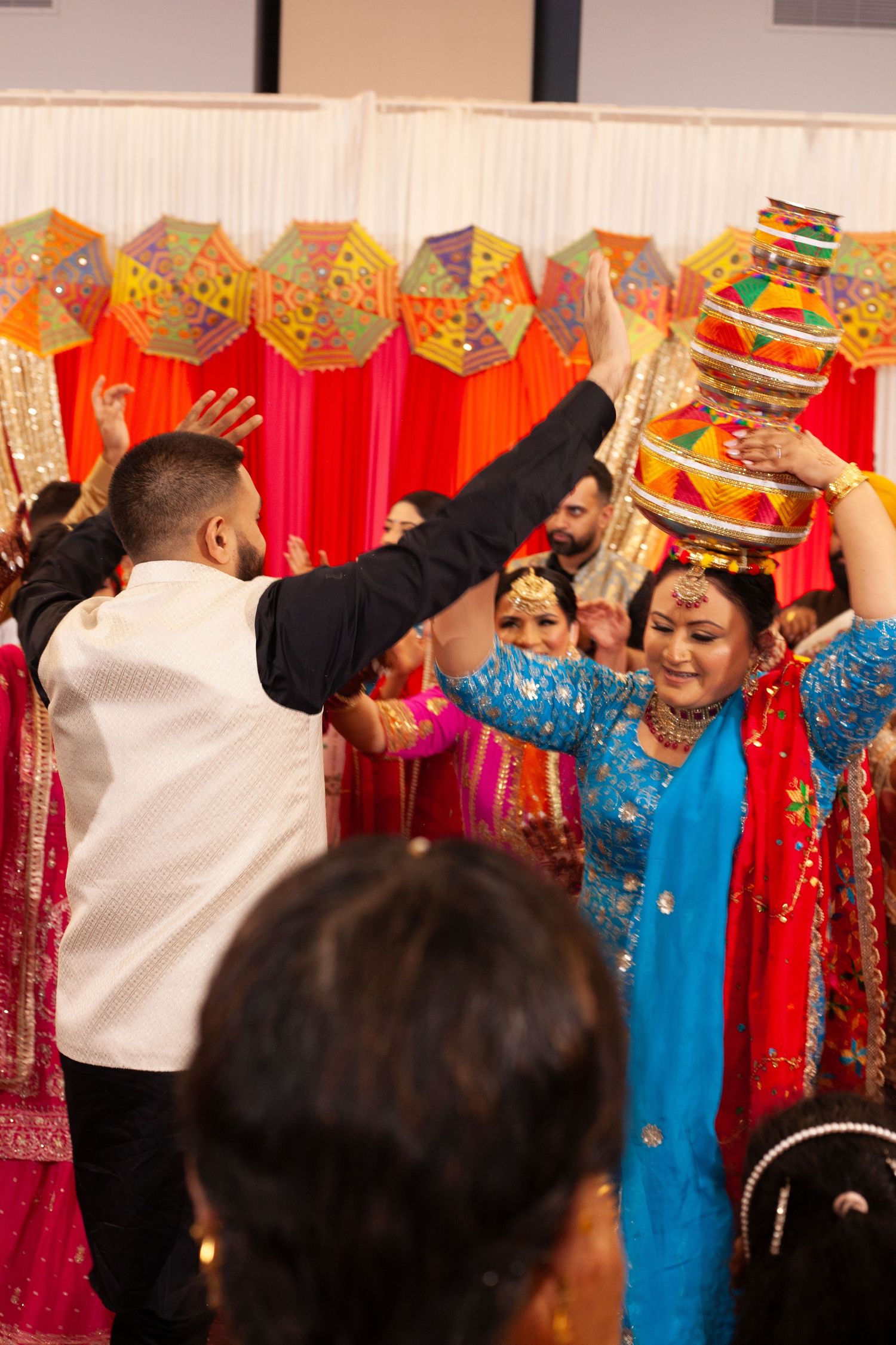 Guests in colorful traditional attire dance with raised arms at a vibrant Punjabi wedding celebration, one woman balancing a decorated pot.