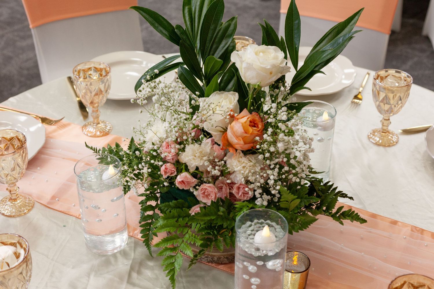 Wedding reception table centerpiece of peach roses, white blooms, baby's breath and greenery with candles, goblets and a blush table runner.