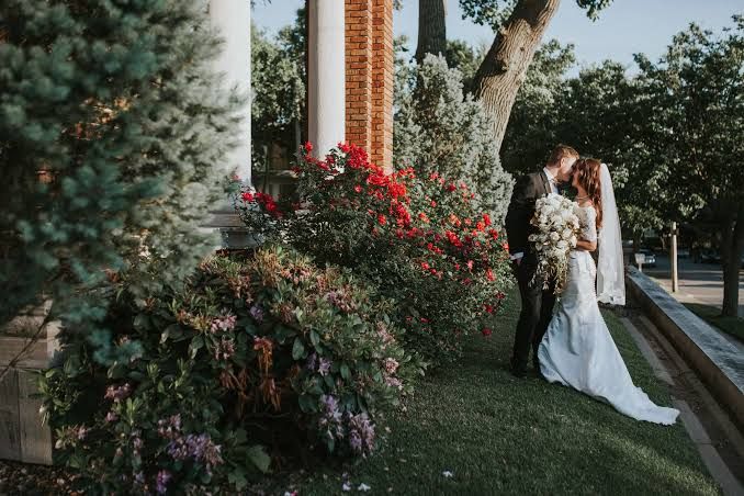 Bride in a white gown and veil embraces her groom on a sunny garden lawn beside red flowering bushes and a brick and white column.