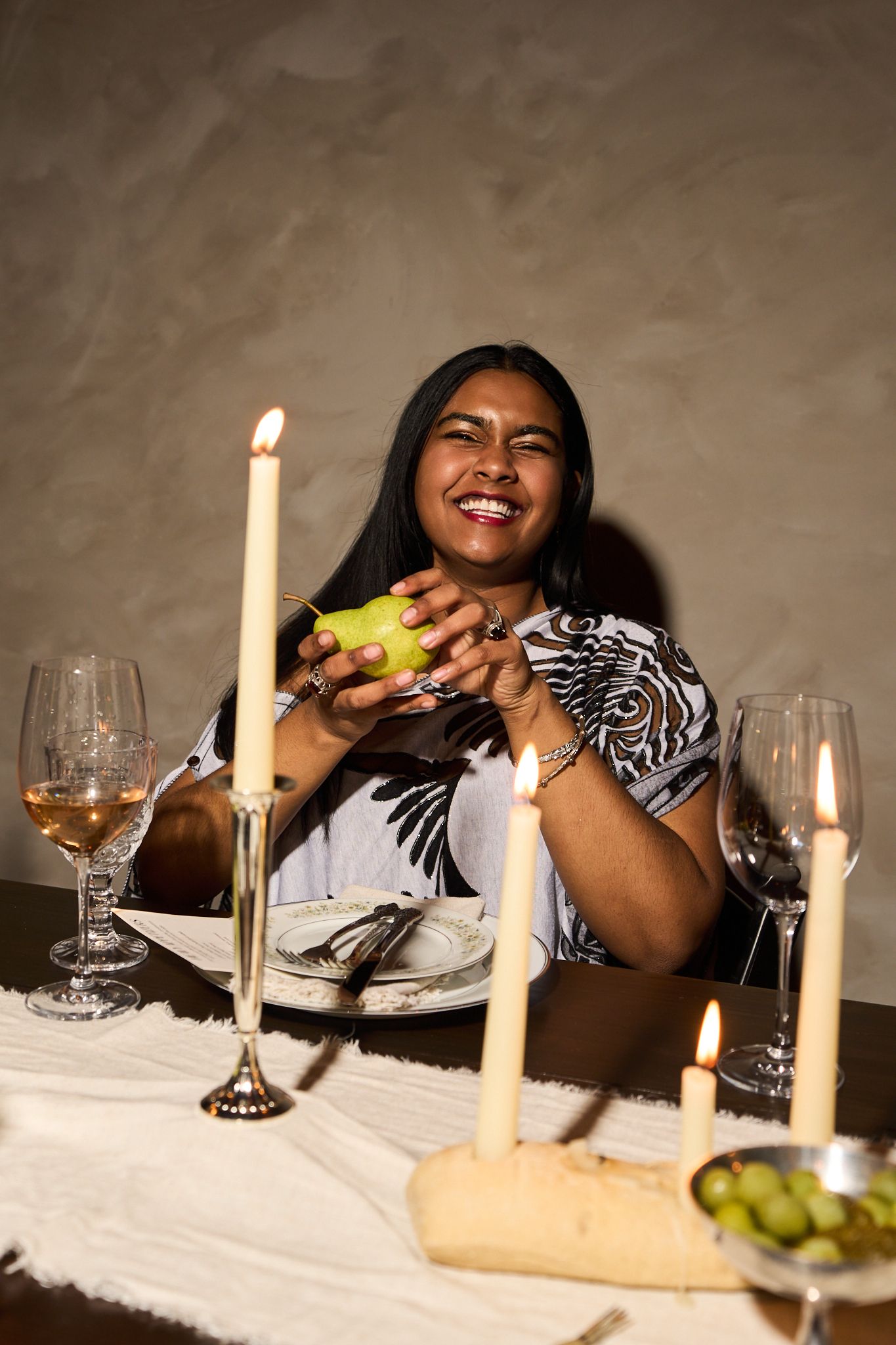 Smiling woman holding a pear at candlelit dinner table with wine glasses, bread, and grapes in elegant setting.