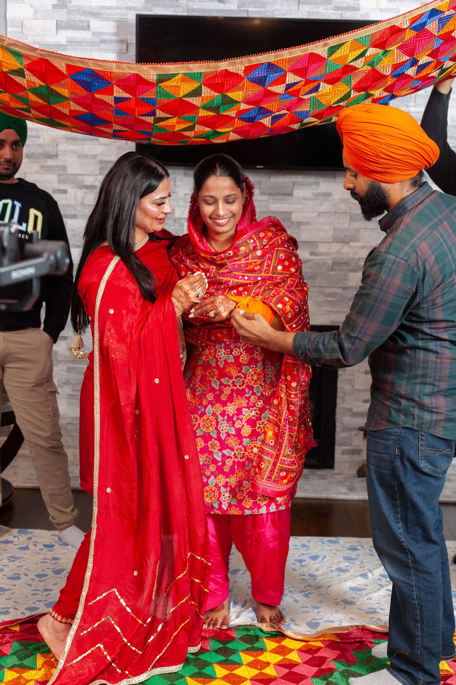 Two women in red traditional Punjabi outfits smile during a ceremony under a colorful phulkari canopy as a man in an orange turban assists.