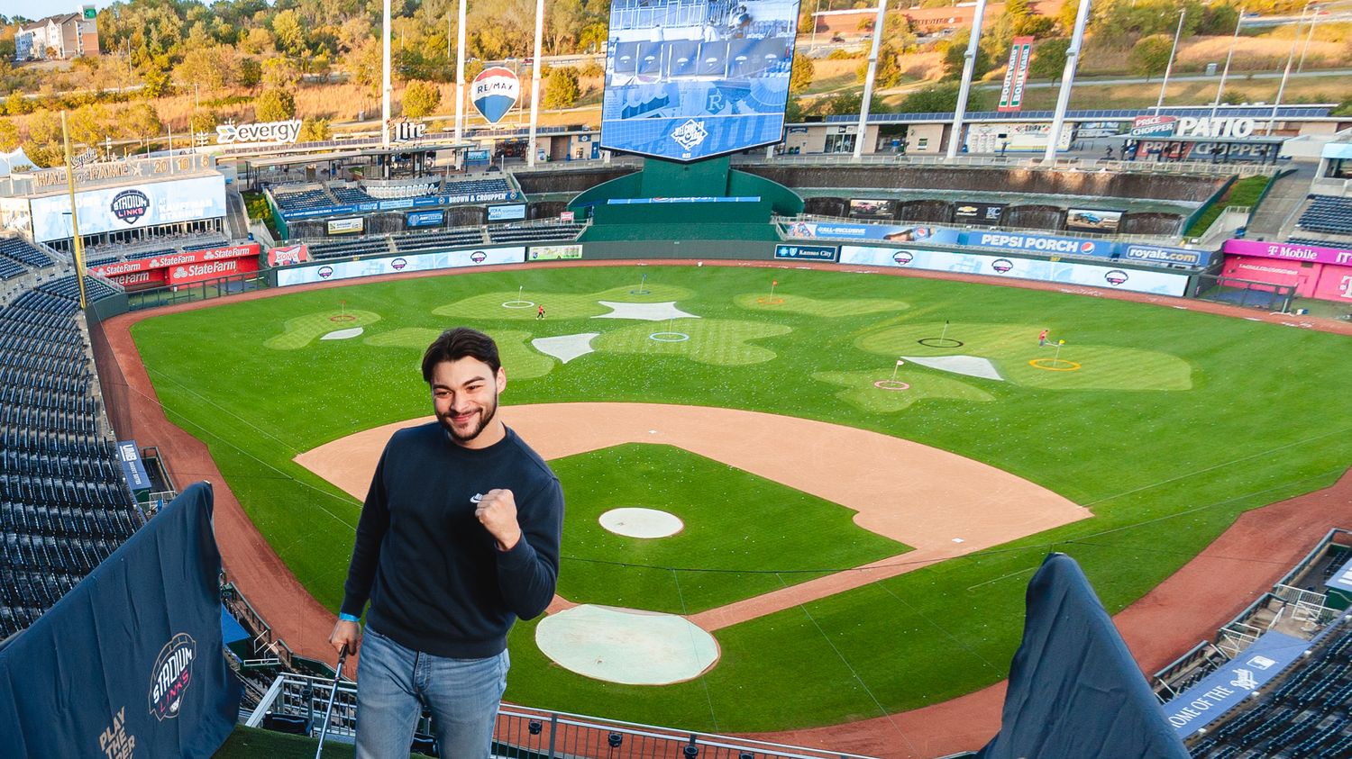 Man celebrating with fist pump at empty baseball stadium with green diamond field and mountain views.