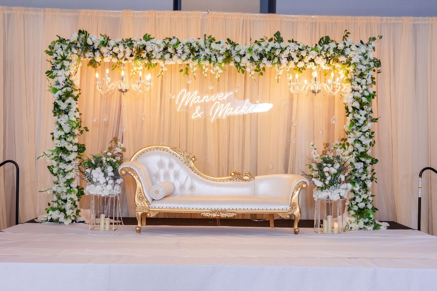Elegant wedding sweetheart stage with a tufted white and gold loveseat, floral arch, warm string lights and a glowing neon name sign.