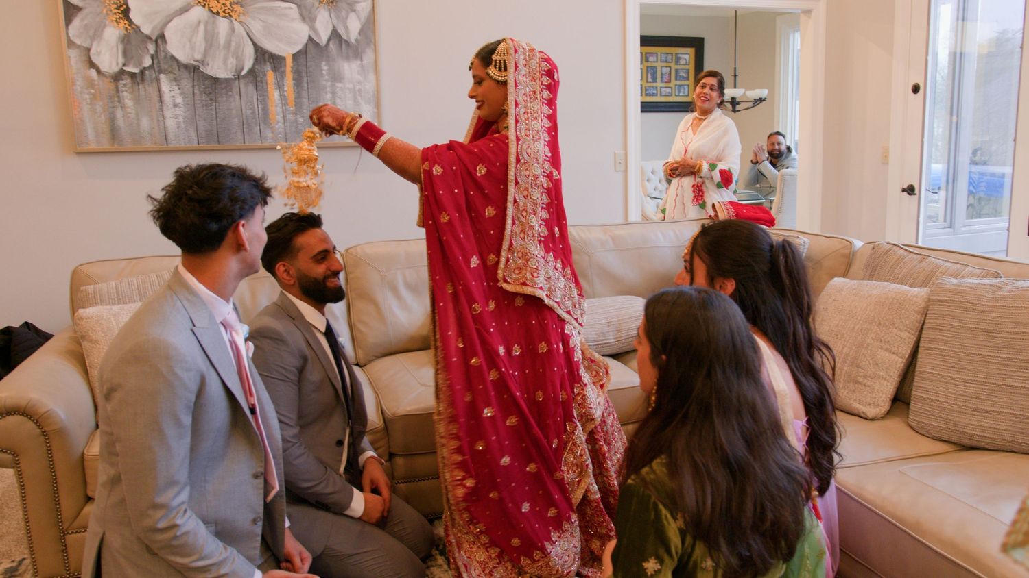 Bride in a red and gold embroidered lehenga adjusts her jewelry while family members sit on a cream sofa in a bright living room.