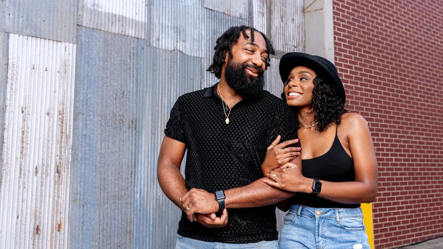 A joyful Black couple embraces and laughs together outdoors near a corrugated metal wall on a sunny day.