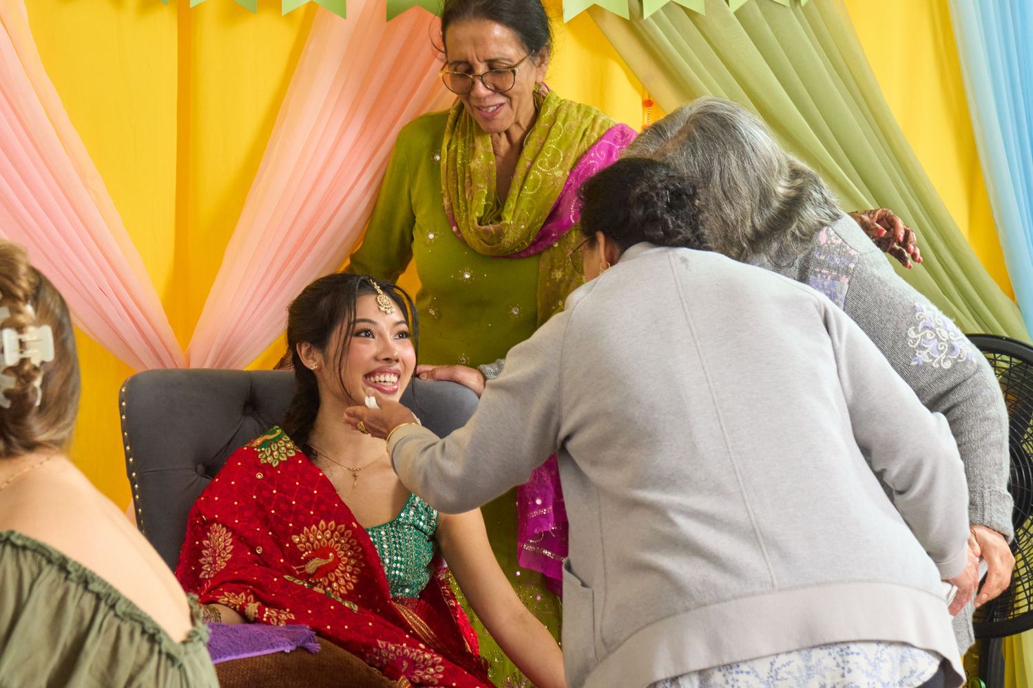 Women in colorful traditional attire celebrate at an Indian ceremony, with an elder blessing a smiling young woman in red.