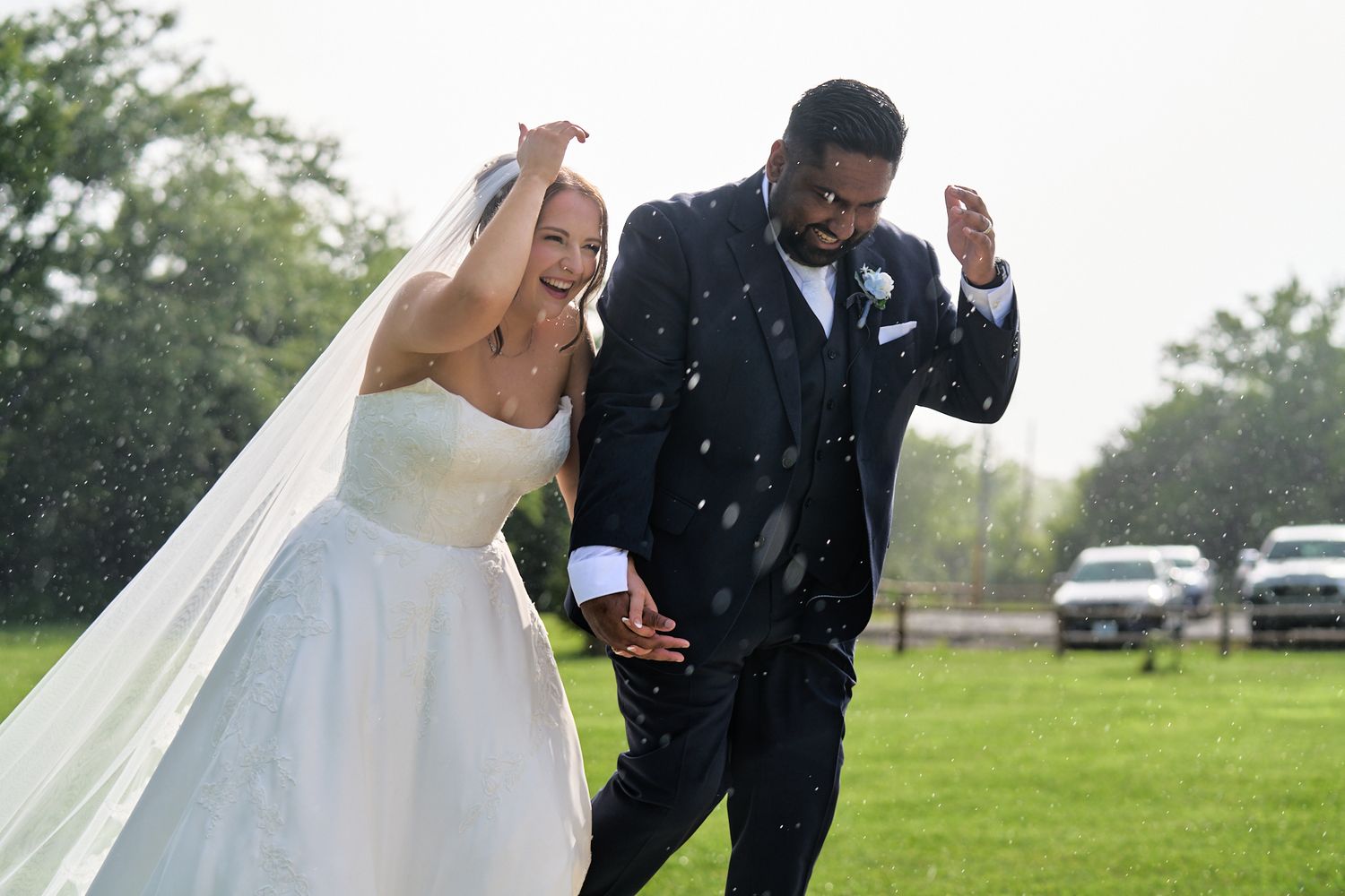Bride in a white gown and veil laughs while holding hands with a groom in a dark suit as rain falls during their outdoor wedding.