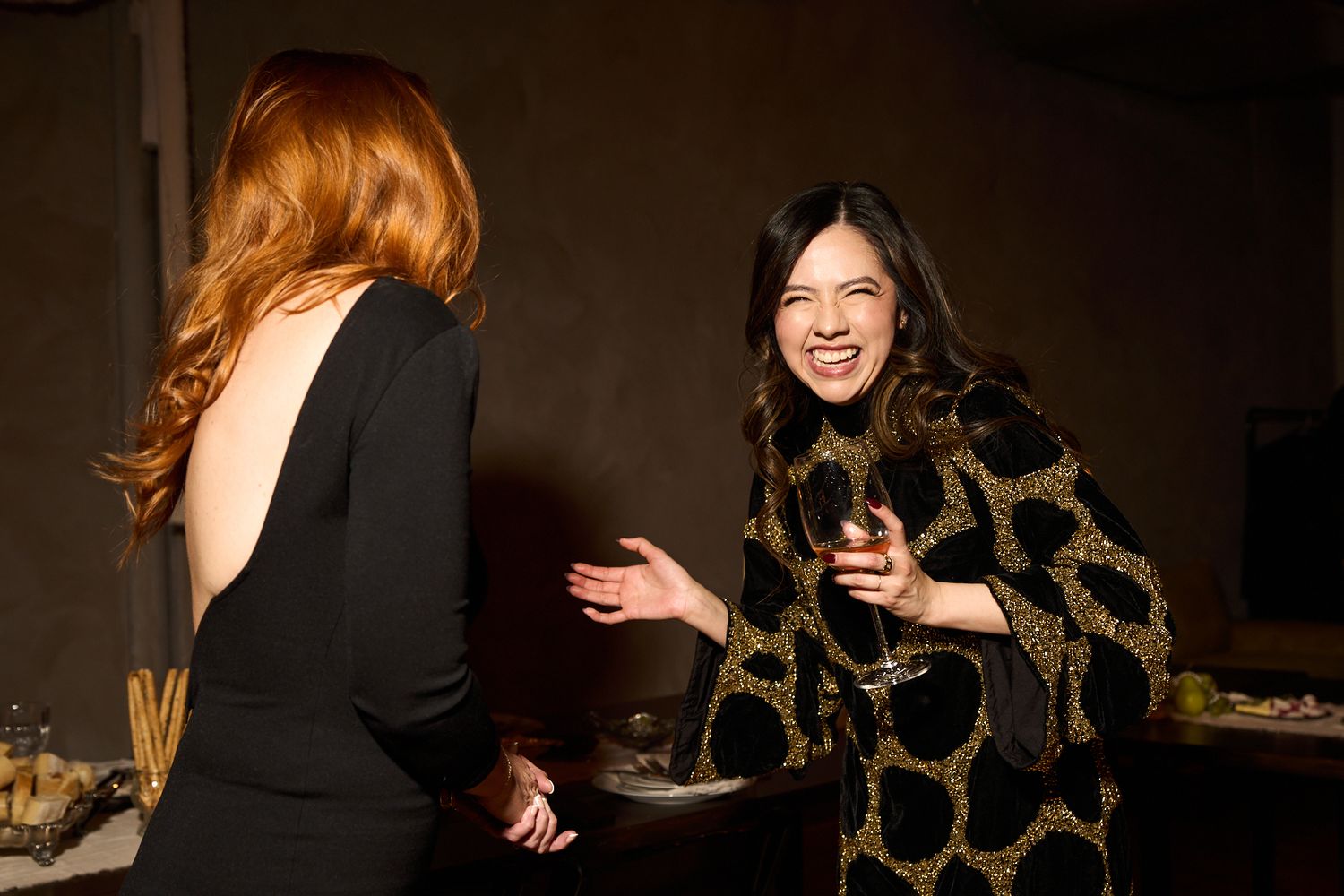 Two women laughing and conversing at an elegant evening event, one in black dress, other in gold patterned coat.