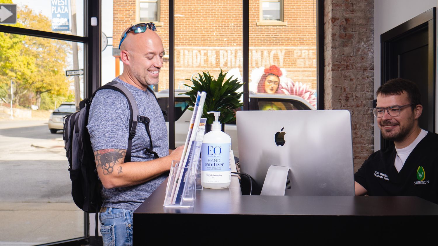 Man with tattoos checks in at a modern dispensary reception desk with hand sanitizer and iMac computer.