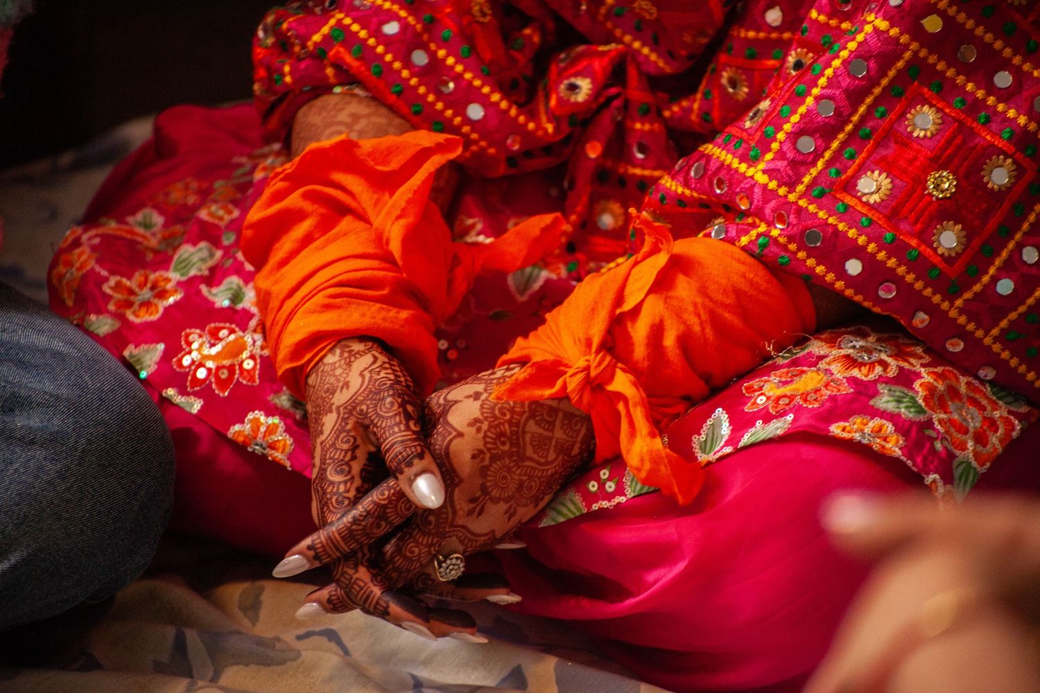 Hands decorated with intricate henna designs rest on vibrant orange and pink embroidered fabrics in a traditional wedding ceremony.
