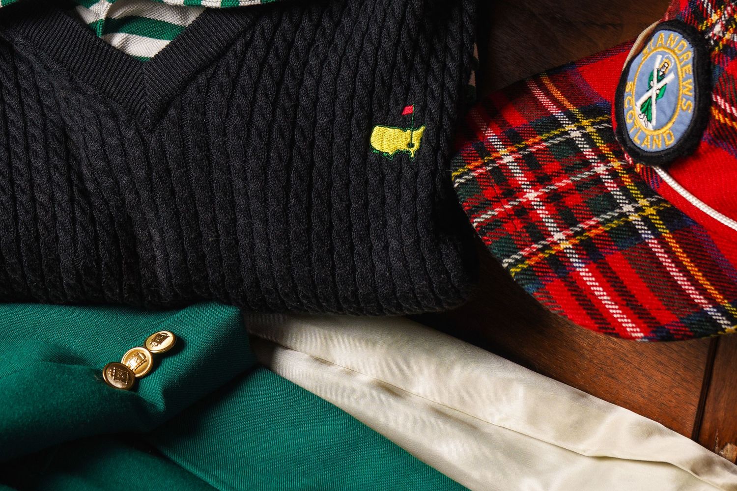 Flat lay of Masters golf black cable knit sweater, green jacket, plaid cap, and white shirt accessories.