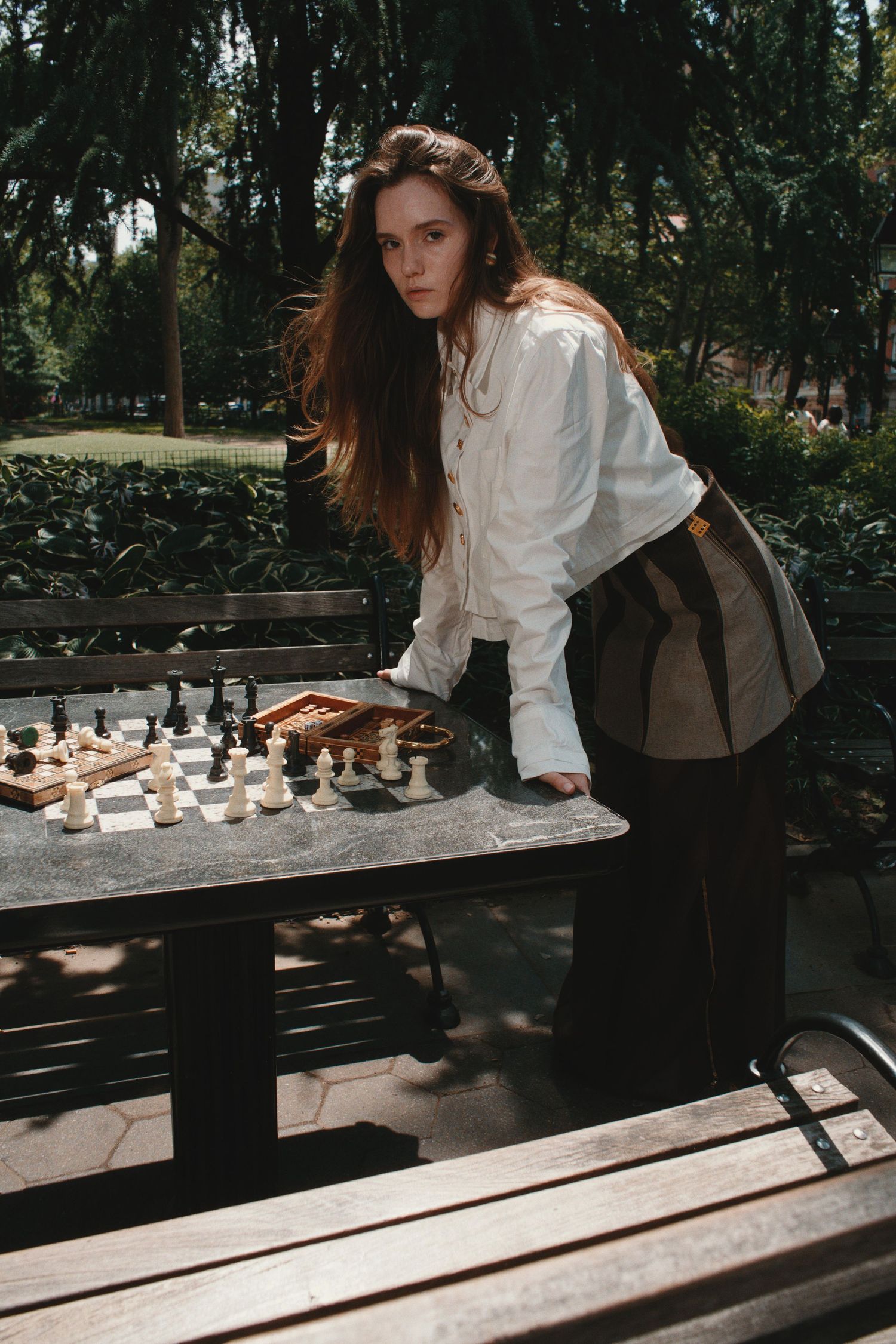 Young woman with long hair leaning over a chess board at an outdoor park table, wearing a white shirt.
