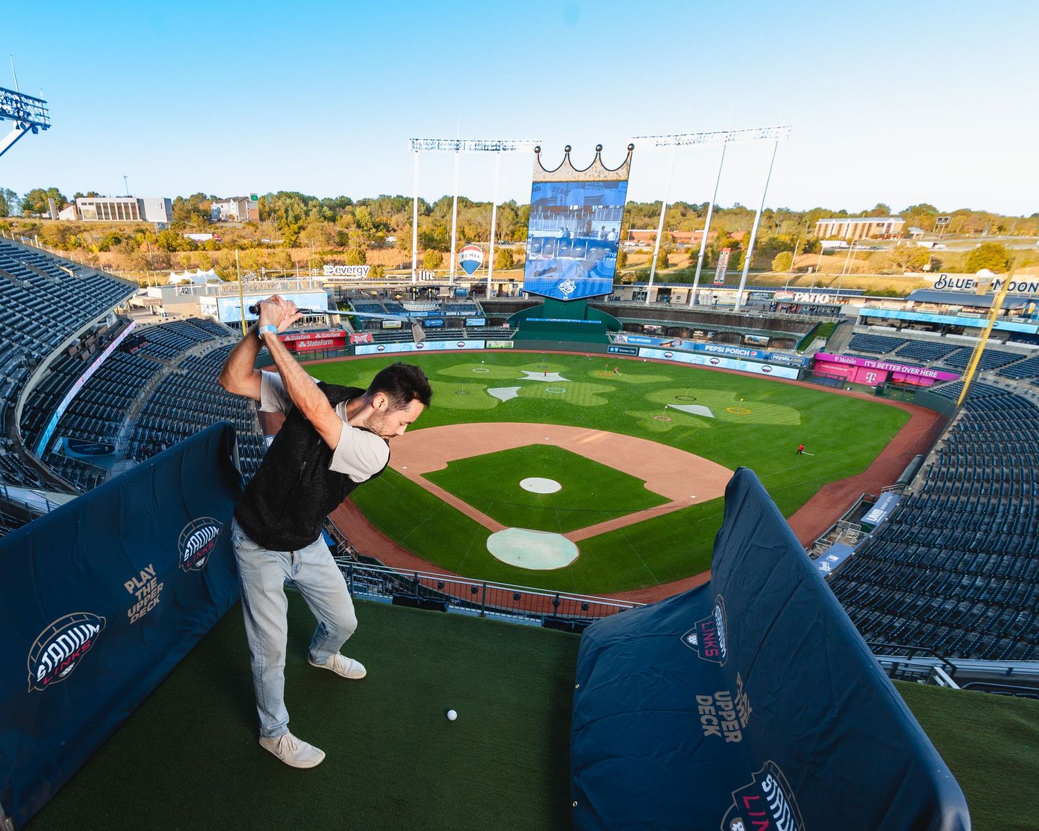 Golfer swinging a club on a tee box set up inside a MLB baseball stadium with green field views.