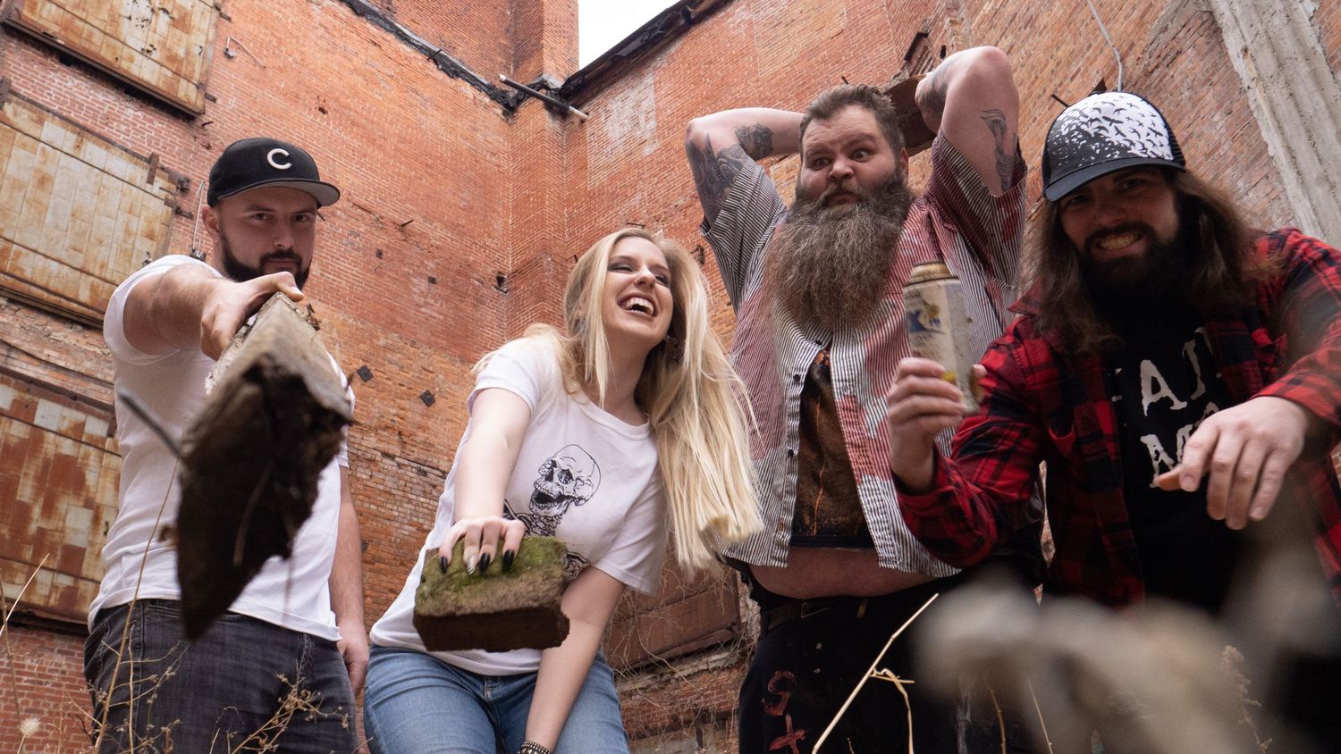 Four friends with beards and long hair laugh outdoors near a brick wall, holding plants and drinks.