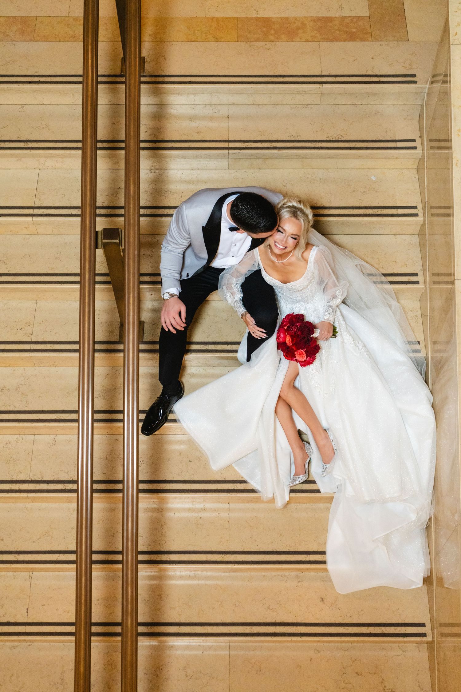 Aerial view of bride and groom sitting on marble stairs with red bouquet.