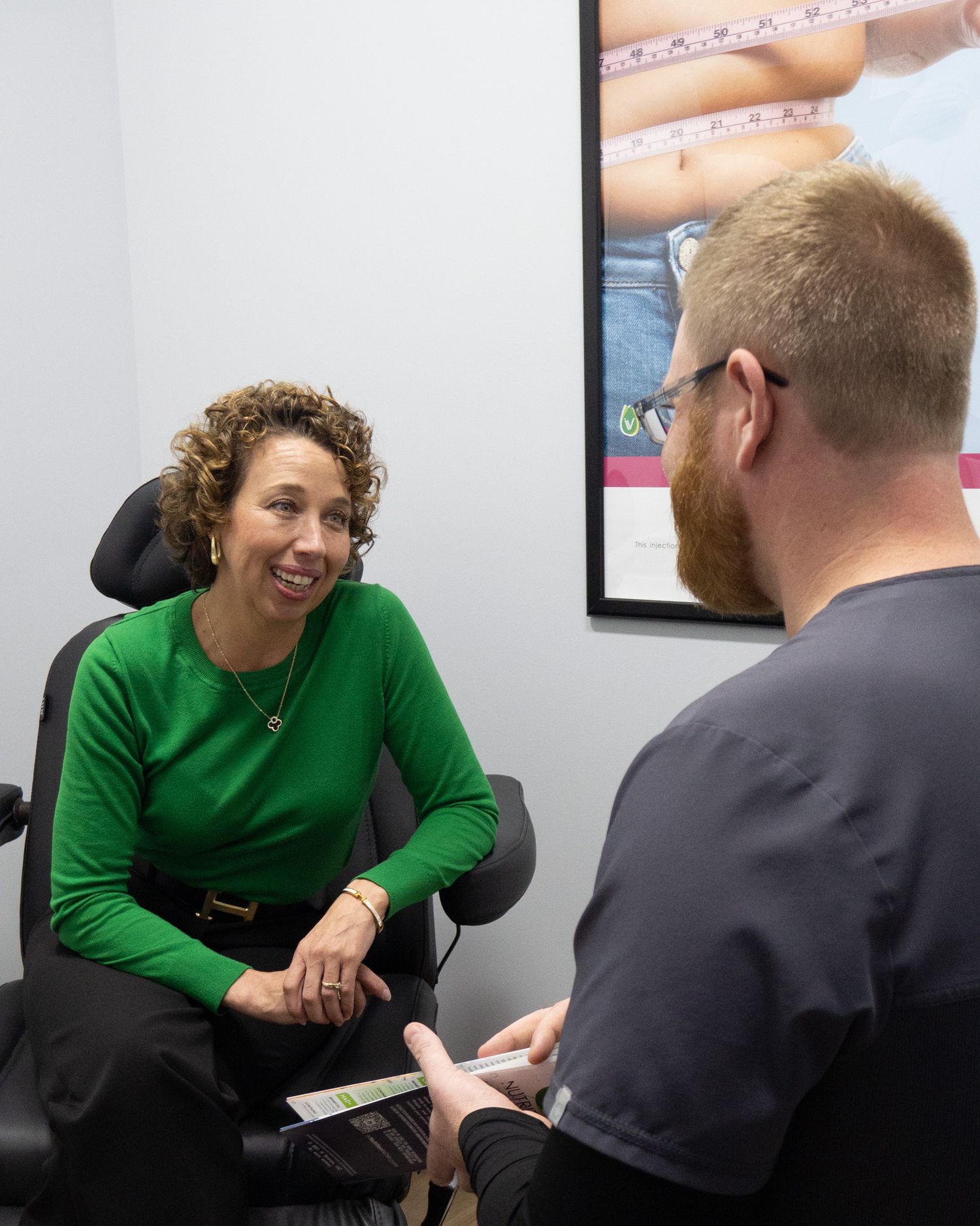 A smiling woman in a green sweater consults with a male healthcare professional in a medical office setting.