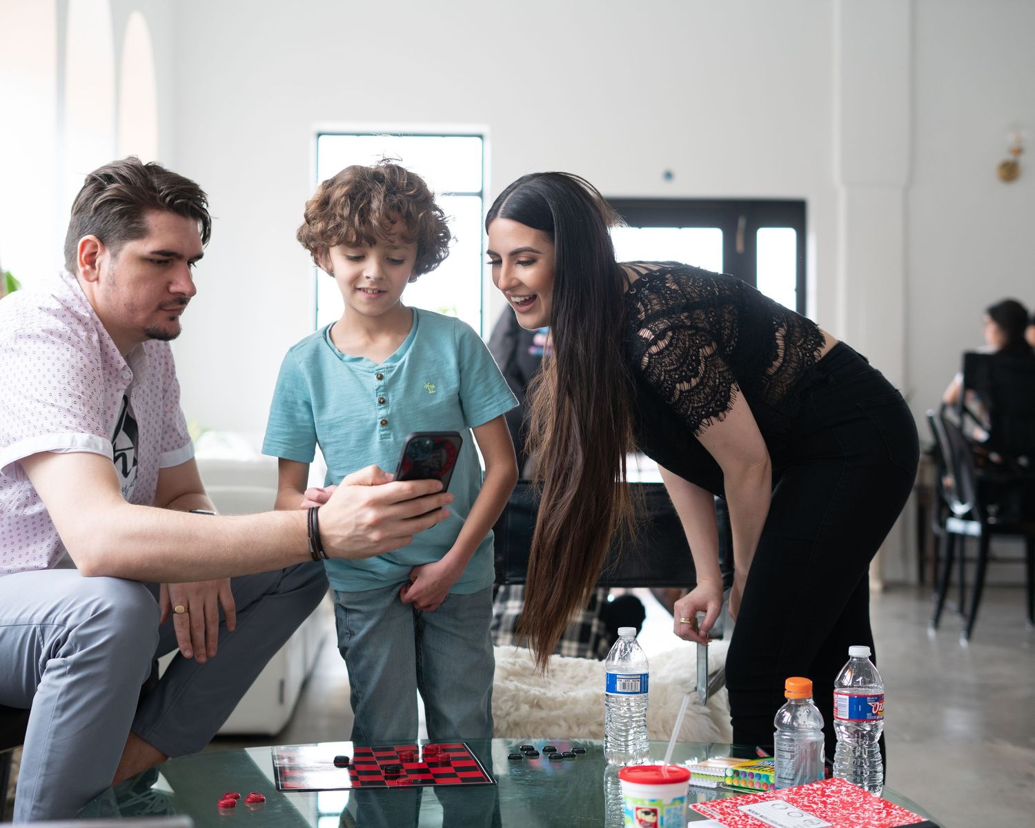 Family of three gathered around a glass table looking at a smartphone together in a bright modern living space.