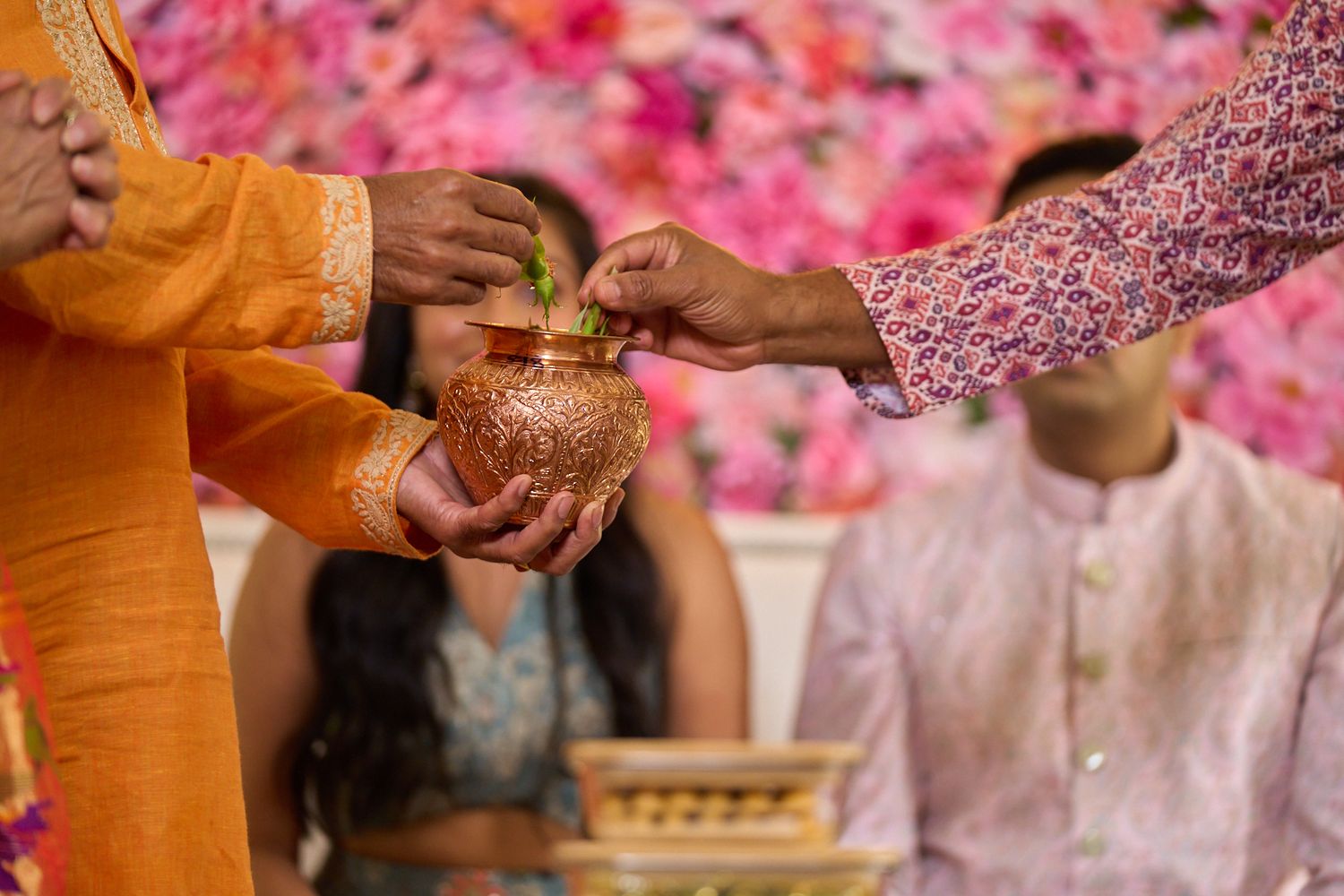 Hands holding a decorated copper kalash with a coconut during a traditional Indian wedding ritual, with guests and pink flowers in the backg