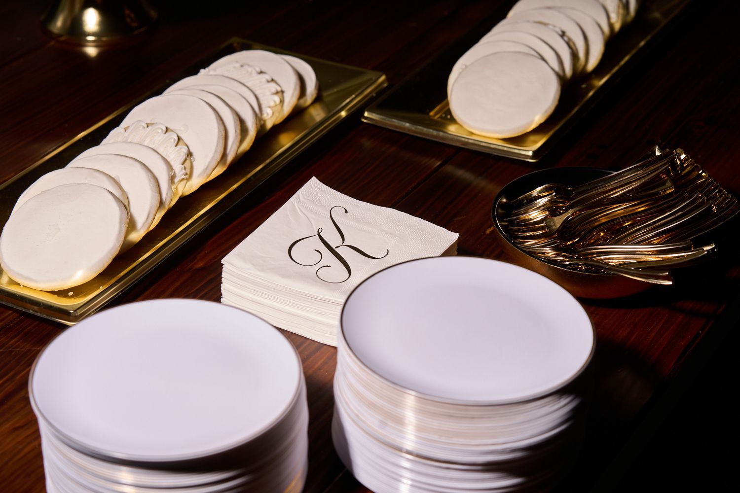 Stacks of white dessert plates beside gold trays of round sugar cookies and a monogrammed K cocktail napkin on a wooden table.