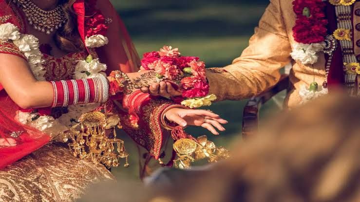 Close-up of a bride and groom's hands joined during a traditional Indian wedding ceremony, adorned with red bangles and floral garlands.