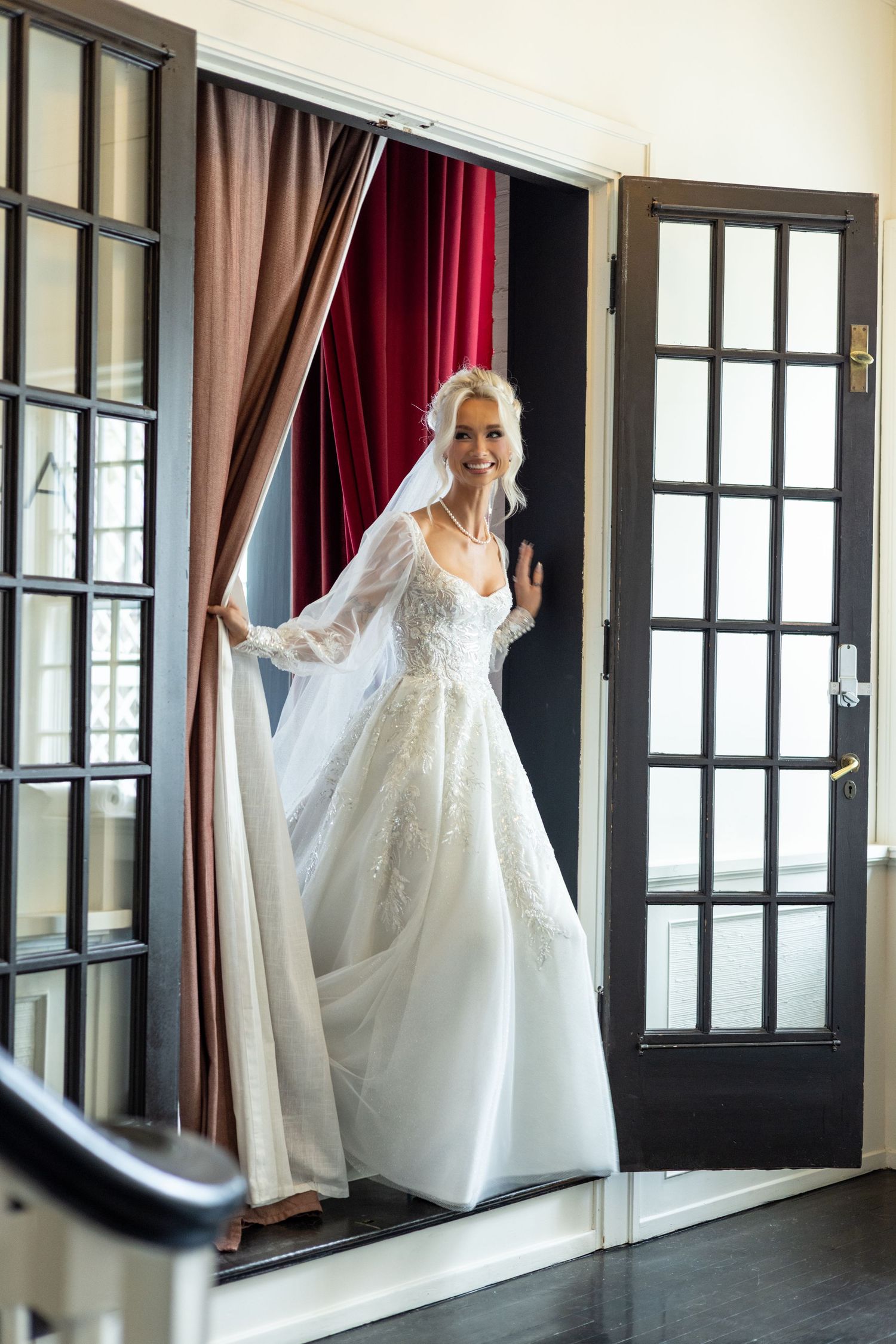 Radiant blonde bride in lace wedding gown with veil standing in doorway with French doors.
