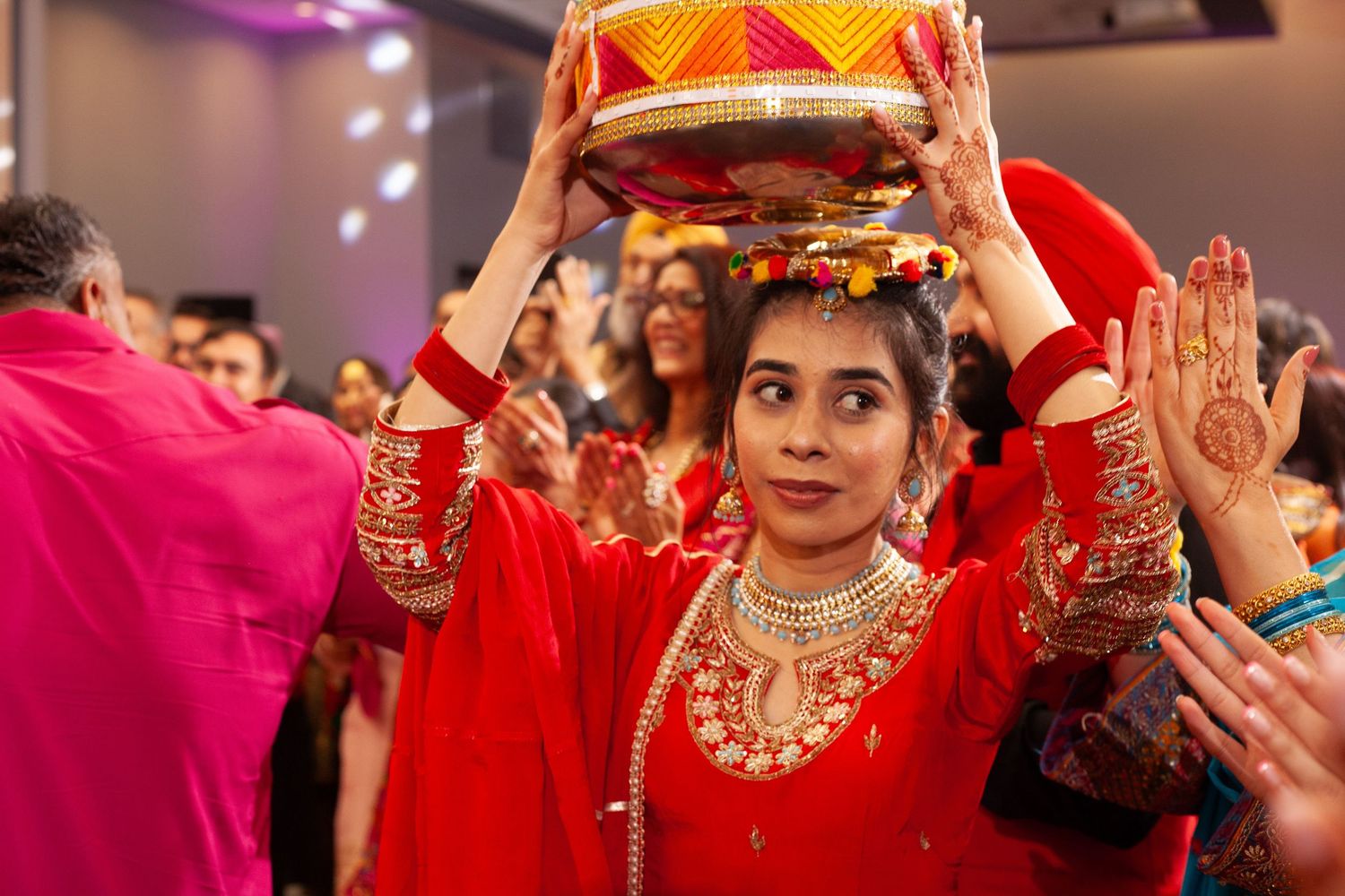 Smiling woman in a red embroidered outfit and gold jewelry balances a decorated pot on her head during a lively traditional celebration.