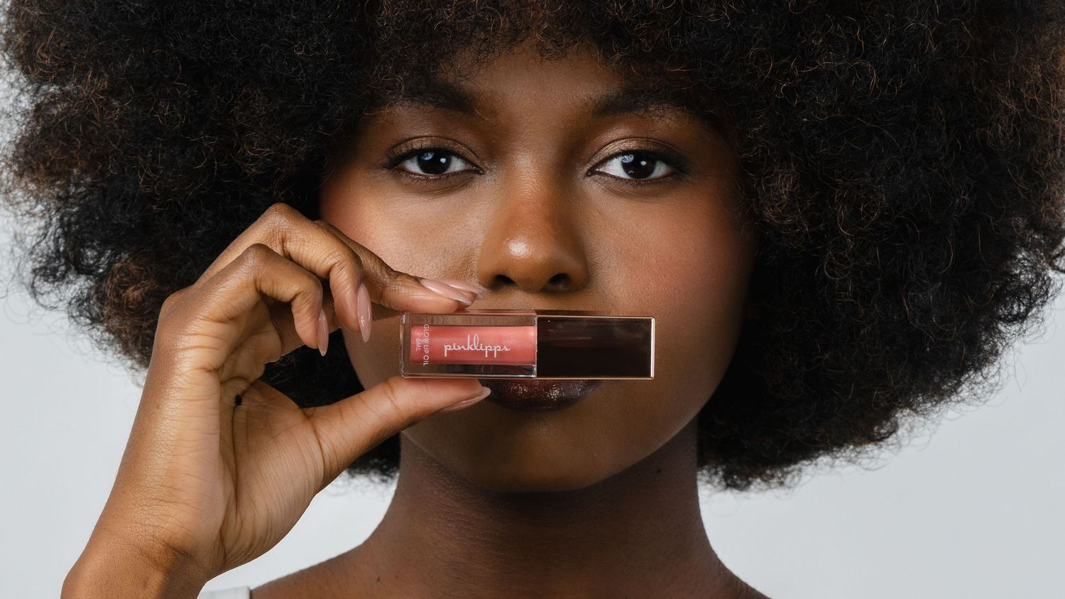 A woman with a natural afro holds a pink lipstick swatch near her lips against a white background.