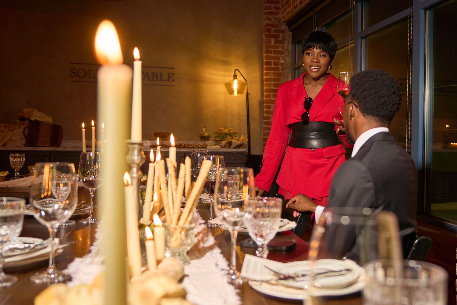 Woman in red dress stands at candlelit dinner table speaking with a seated guest in elegant restaurant.