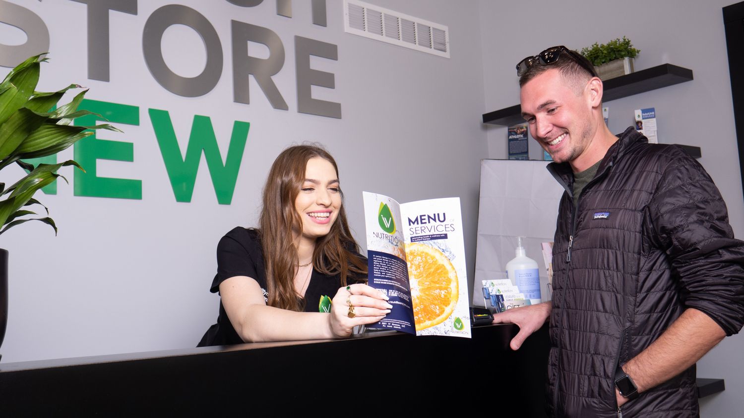 Smiling dispensary staff member shows menu to happy male customer at a modern cannabis store reception desk.
