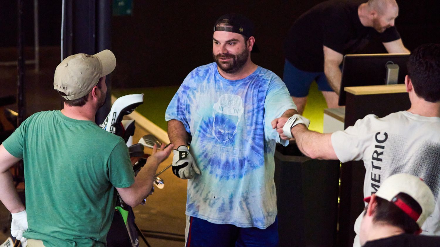 Man in blue tie-dye shirt gestures on film set surrounded by crew members holding equipment.