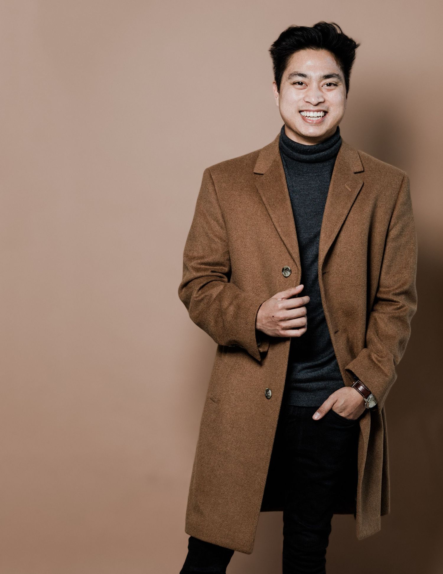 Smiling Asian man in camel wool overcoat and gray turtleneck posing against warm brown background.