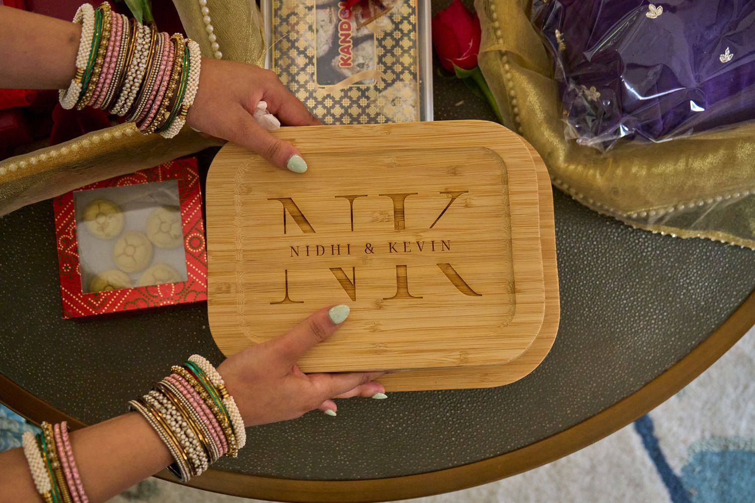 Personalized engraved bamboo cutting board with monogram NK for Nighi and Kevin, held by hands wearing colorful bangles.