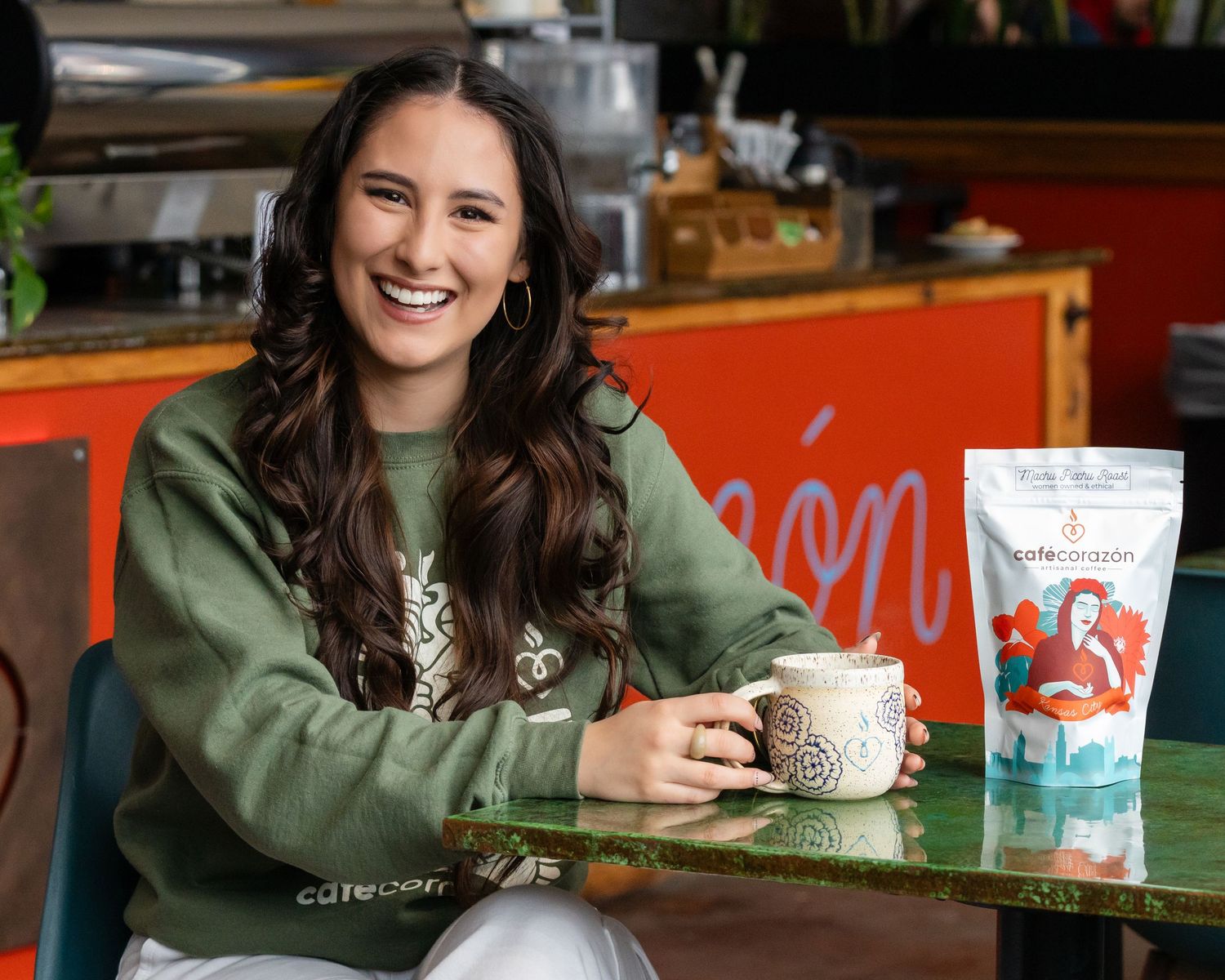 Smiling woman in green sweatshirt holding ceramic mug beside branded coffee bag at colorful cafe table.
