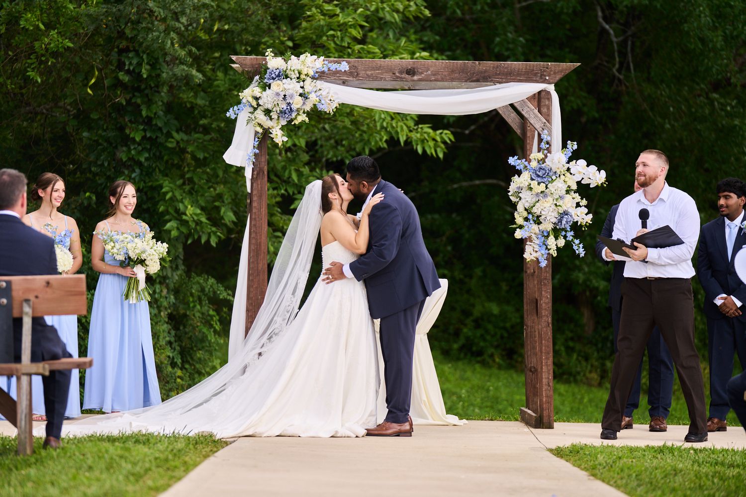 Bride and groom kiss under a rustic wooden wedding arch with white drapes and blue floral accents as guests and an officiant watch outdoors.