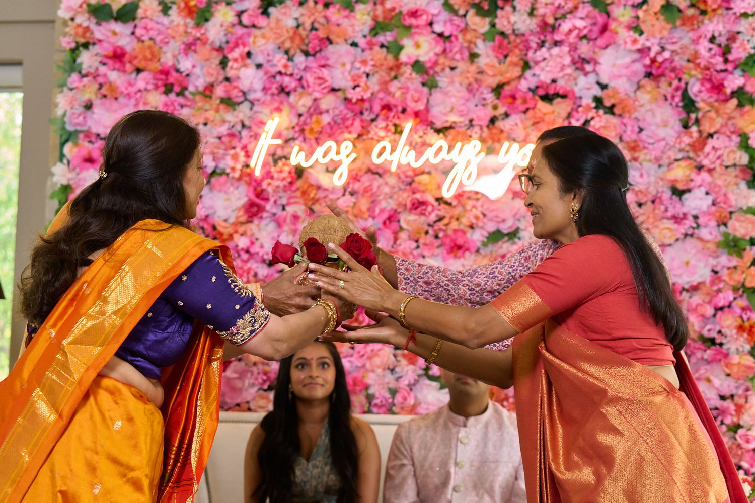 Two women in orange and coral sarees exchange a ceremonial gesture before a pink floral wall with a neon sign at an Indian celebration.