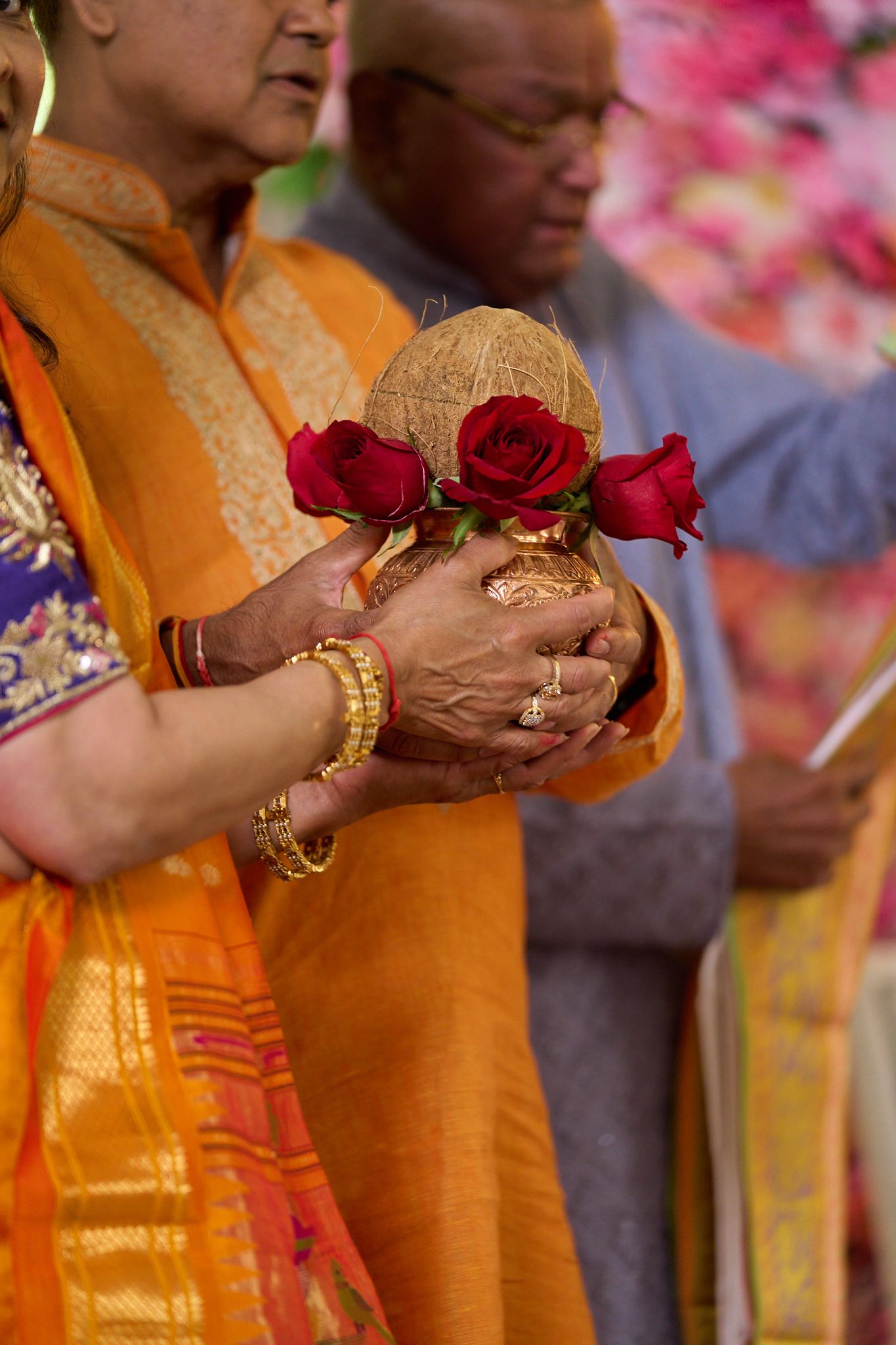 Indian woman in orange saree holding a kalash with red roses during a Hindu ceremony.
