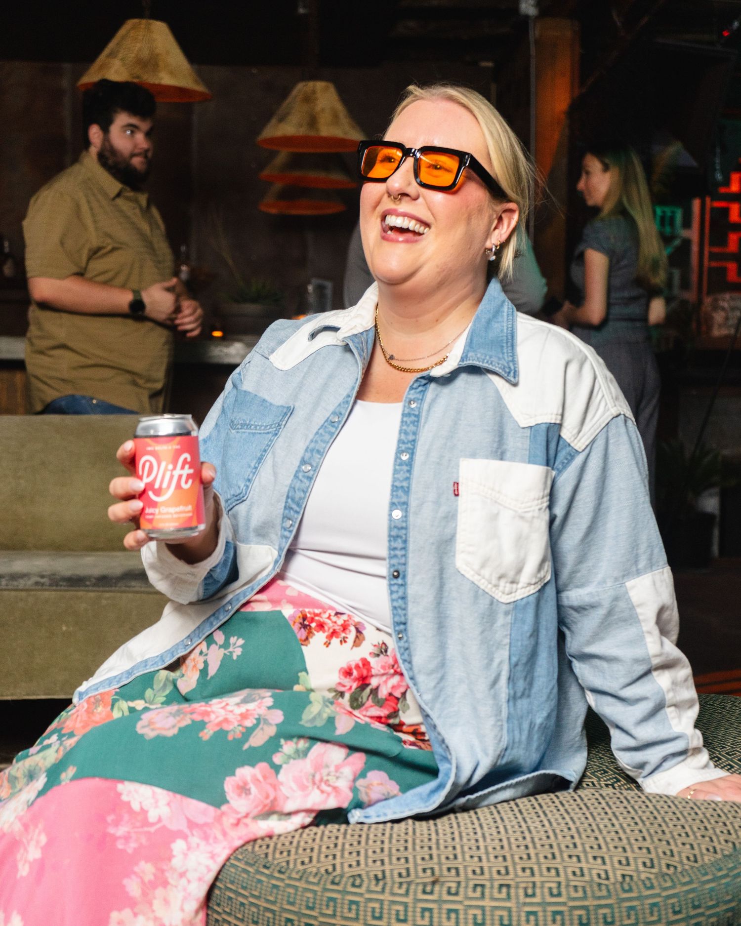 Laughing blonde woman in denim shirt and floral skirt holding a Olipop soda can, wearing orange sunglasses outdoors.