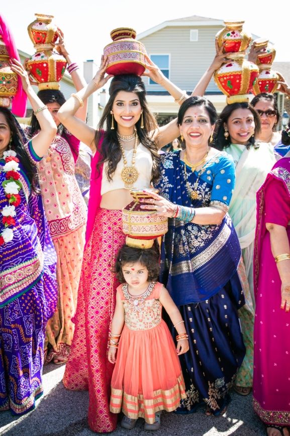 Smiling women in colorful traditional Indian attire carry decorated ceremonial pots on their heads while a little girl in orange joins them.