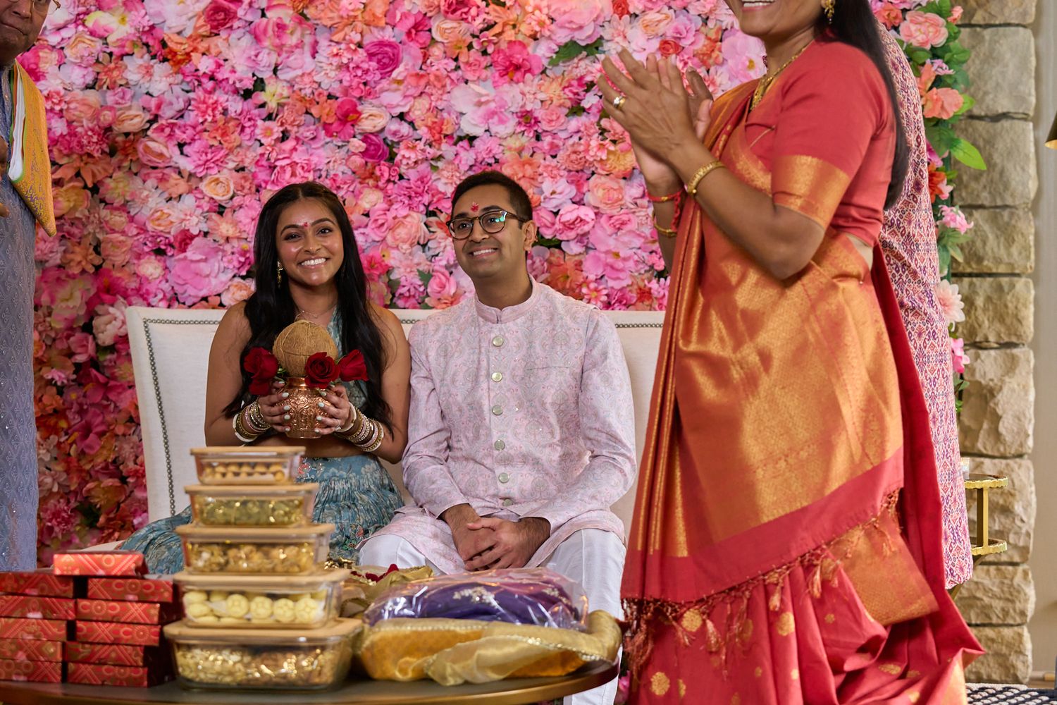 A smiling couple in traditional Indian attire sits before a pink floral wall as a woman in an orange silk saree stands near gift boxes.