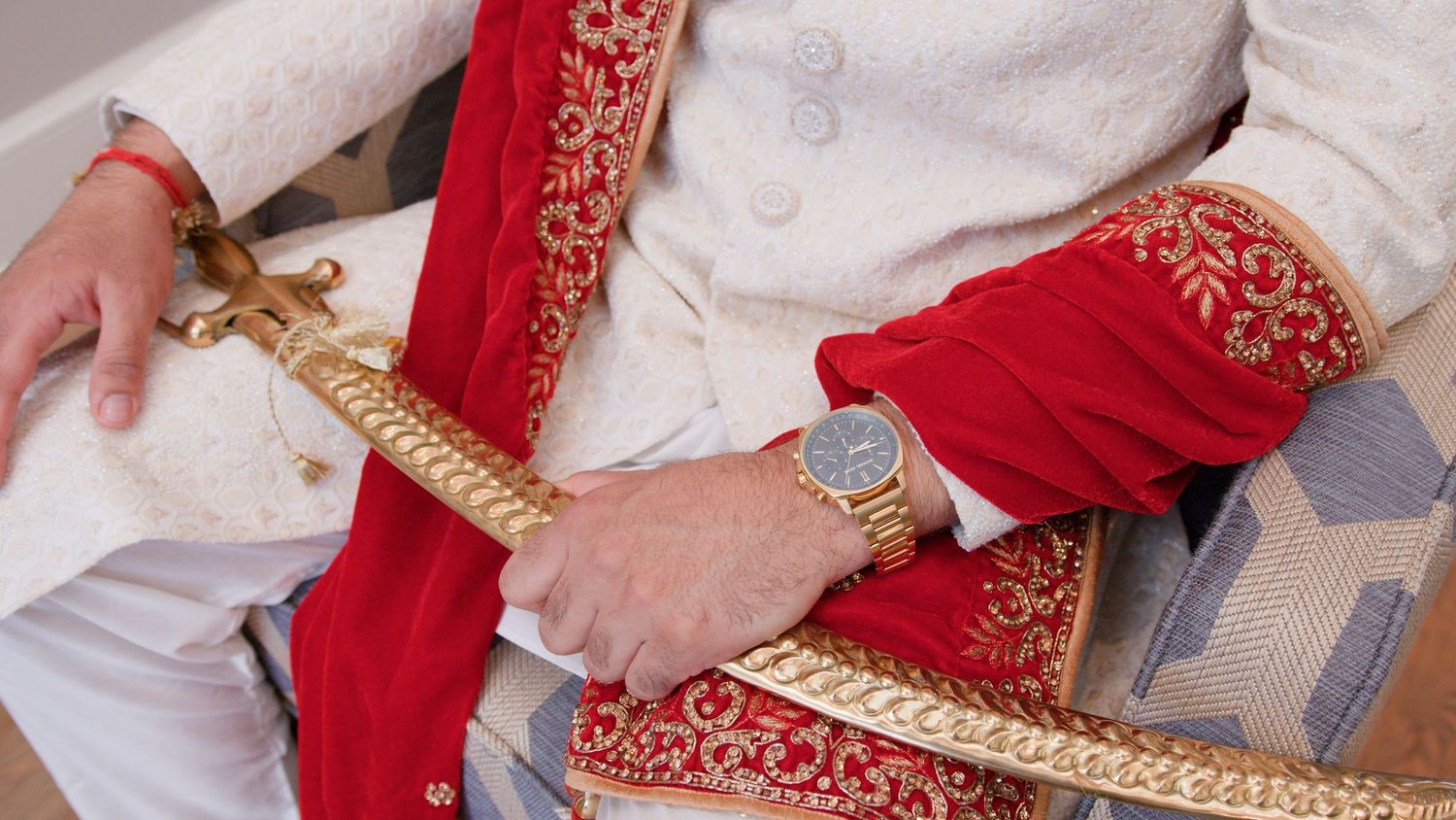 Groom in an ivory sherwani with red embroidered shawl rests his hands on a golden ceremonial sword, wearing a gold wristwatch.