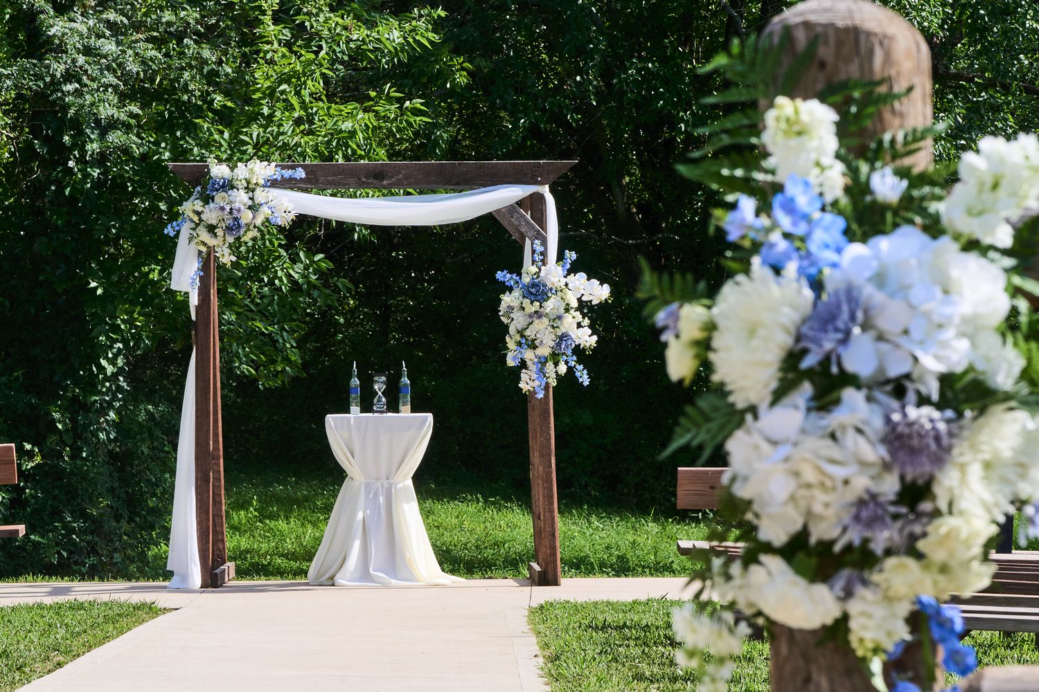 Outdoor wedding ceremony arch with white draped fabric and blue and white floral arrangements beside a small table in a green garden.