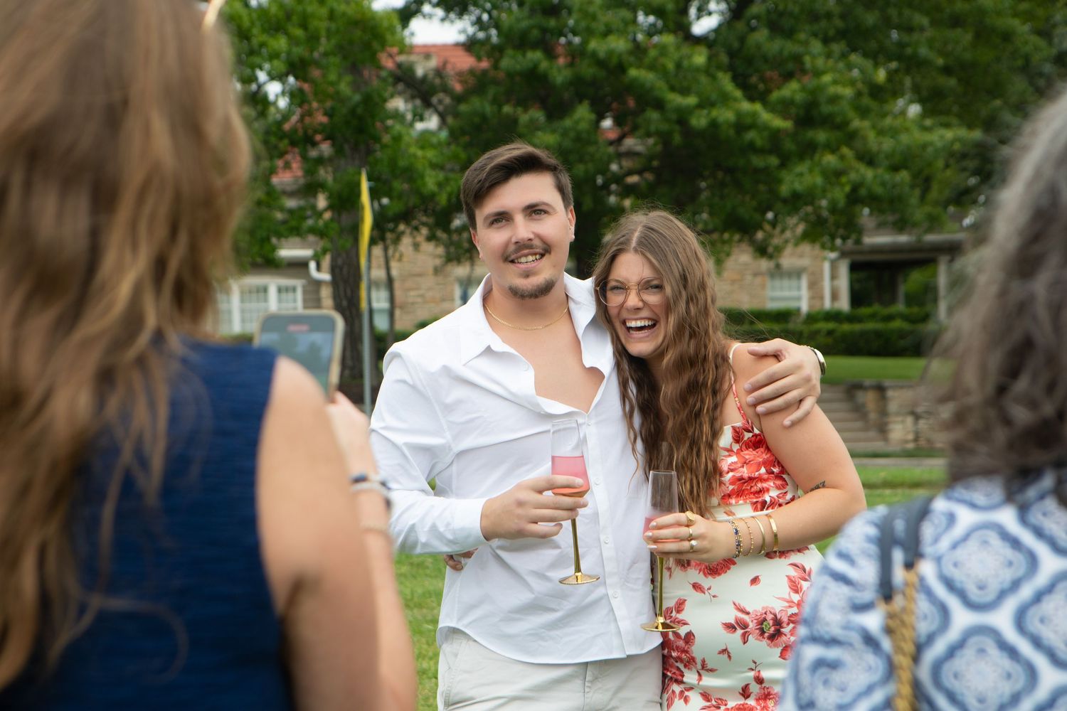 A smiling couple poses outdoors at a garden party, man in white shirt holding a pink drink, woman in floral dress.
