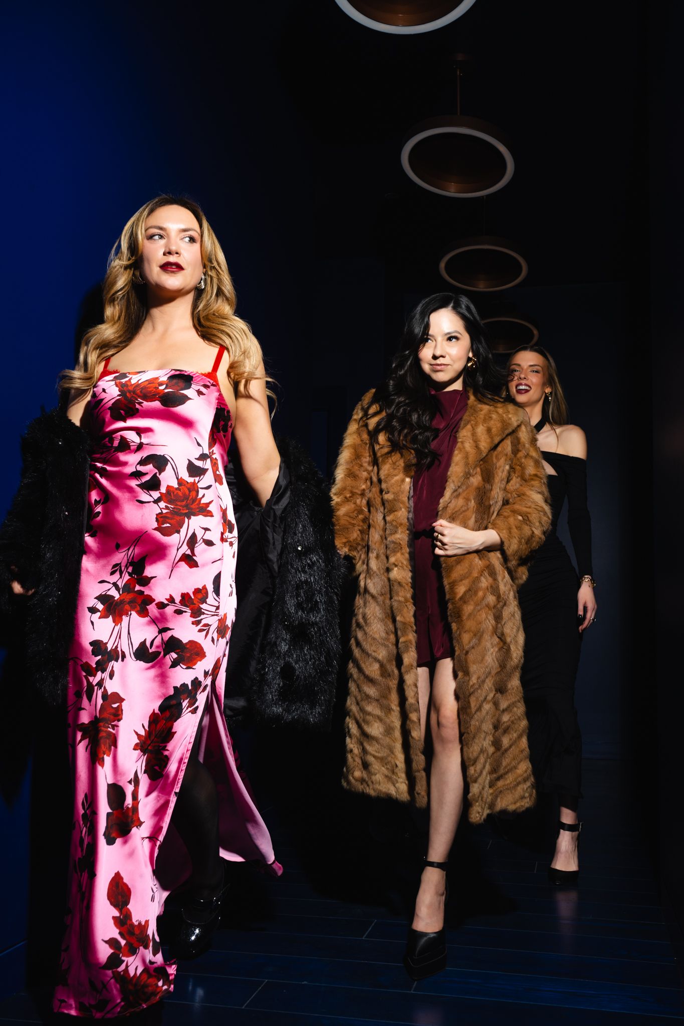 Three women walk a runway, wearing a floral gown, fur coat, and black dress at a fashion show.