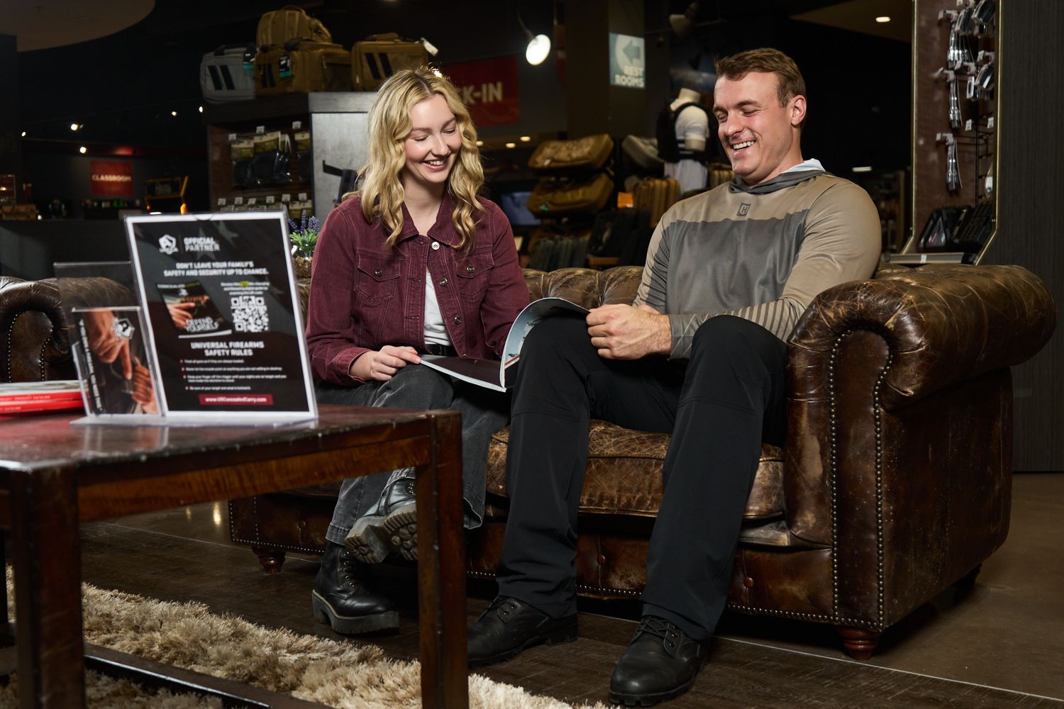 A man and woman sit on a leather couch in a store, smiling and reading a magazine together.